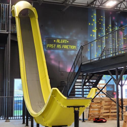 A bright yellow slide in a modern indoor space, with a sign reading "ALERT: FAST AS FRICTION" above it.