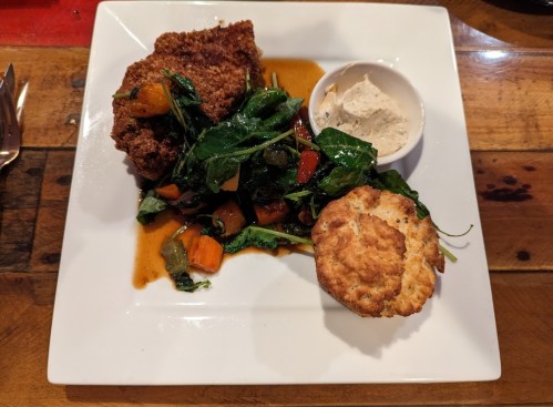 A plate featuring fried chicken, sautéed greens with vegetables, and a biscuit, served with a small bowl of sauce.