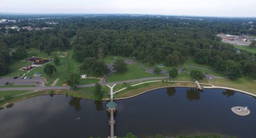 Aerial view of a park with a pond, walking paths, and green trees, featuring playgrounds and parking areas nearby.