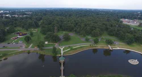 Aerial view of a park with a pond, walking paths, and green trees, featuring playgrounds and parking areas nearby.