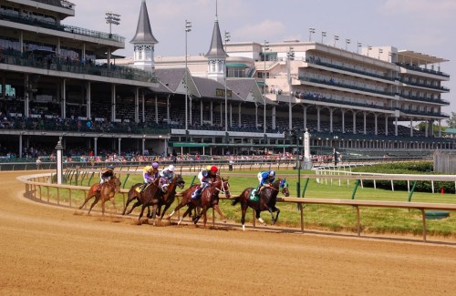 A horse race in progress at a large racetrack with spectators in the stands and a grand building in the background.