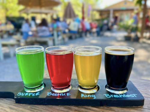 A flight of four colorful drinks labeled "Supper," "Beetnik," "King," and "Breakfast" on a wooden paddle.