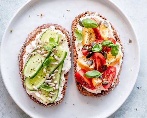 Two open-faced sandwiches on a white plate: one topped with cucumber and sunflower seeds, the other with tomatoes and peppers.