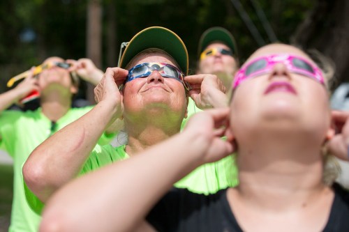 A group of people wearing colorful sunglasses gazes upward, enjoying a solar event.