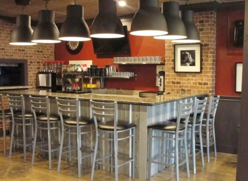 A modern bar with a granite countertop, metal stools, and pendant lights, set against a brick wall.