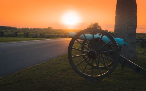 A historic cannon sits beside a road at sunset, with a vibrant orange sky and silhouettes of trees in the background.
