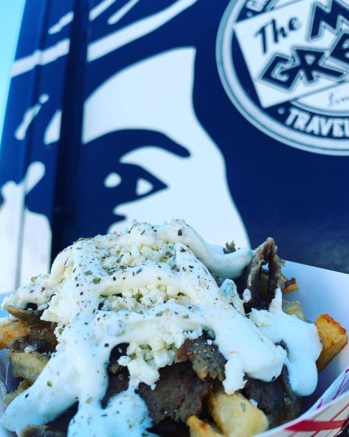 A close-up of a loaded plate of fries topped with meat, white sauce, and crumbled cheese, with a food truck in the background.
