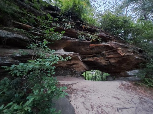 A natural rock formation with an arch, surrounded by lush greenery and a sandy path leading through it.