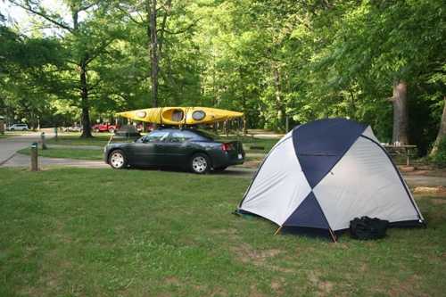 A black car with a yellow kayak on top parked near a tent in a green, wooded campsite.