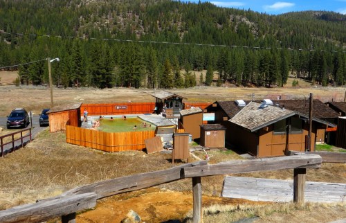 Aerial view of a rustic lodge area with cabins, a pool, and surrounding pine trees under a clear blue sky.