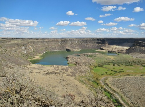 A vast canyon landscape with a winding river, green patches, and scattered clouds in a blue sky.