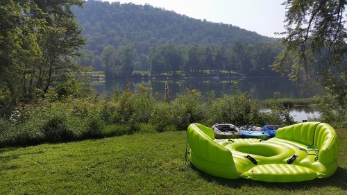 A bright green inflatable raft rests on grass by a calm river, surrounded by trees and a scenic hillside.