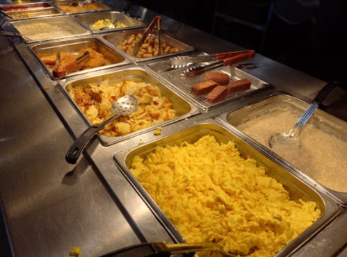 A buffet spread featuring scrambled eggs, hash browns, sausage, and grits in metal trays with serving utensils.