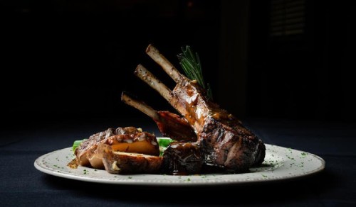 A beautifully plated rack of lamb with sauce, accompanied by roasted potatoes and green vegetables on a dark background.