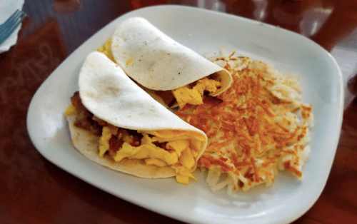 Two breakfast tacos filled with scrambled eggs and meat, served with a side of crispy hash browns on a white plate.