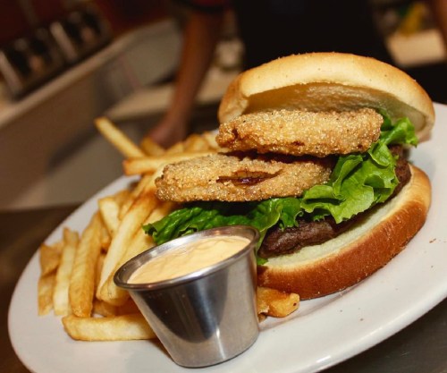 A burger with lettuce and fried onion rings, served with fries and a small cup of dipping sauce on a plate.