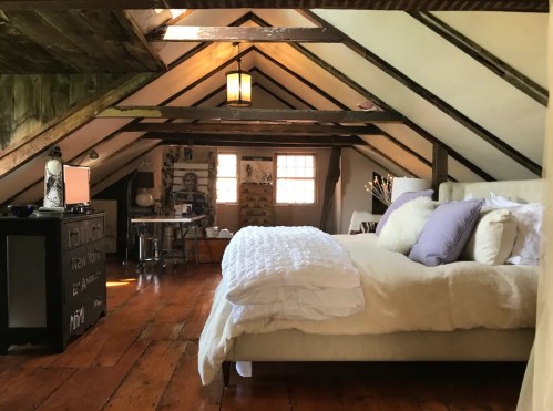 Cozy attic bedroom with a large bed, wooden floors, and a workspace, featuring sloped ceilings and warm lighting.