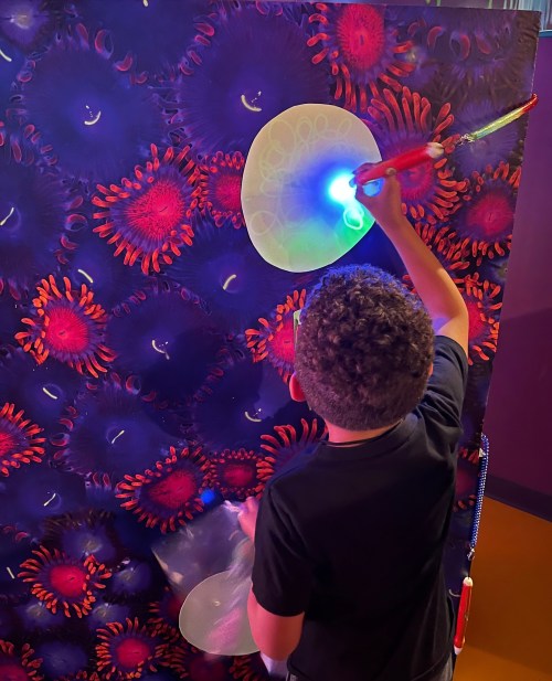 A child uses a light pen to draw on a colorful, textured wall featuring jellyfish and coral designs.