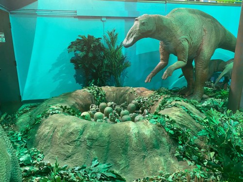 A dinosaur stands near a nest of eggs surrounded by lush greenery in a vibrant, colorful setting.