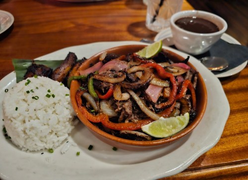 A vibrant dish featuring grilled meat with peppers and onions, served with rice, lime wedges, and a side of sauce.