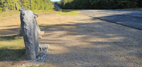 A weathered bench beside a gravel path, with a road and trees in the background under a clear sky.