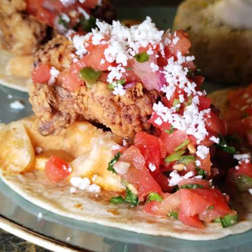 Close-up of tacos topped with fried chicken, fresh tomato salsa, and crumbled cheese on a decorative plate.