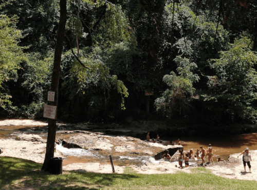 A serene river scene with people enjoying the water, surrounded by lush green trees and rocky banks.
