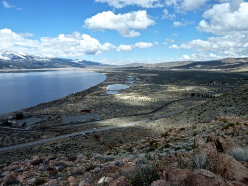 A scenic view of a lake surrounded by hills and clouds, with patches of sunlight illuminating the landscape.