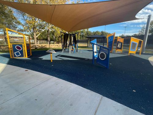 Colorful playground equipment with various shapes and textures under a shaded area, surrounded by trees.