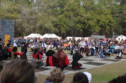A crowd watches a medieval performance in a forested area, featuring actors in period costumes on a stone stage.