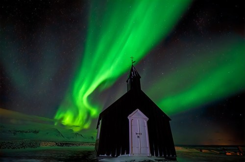 A small black church silhouetted against a vibrant green aurora borealis in a starry night sky.