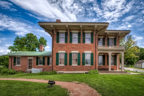 Historic brick house with green shutters, surrounded by grass and trees, under a partly cloudy sky.