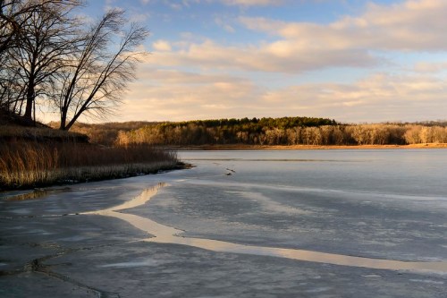 A serene winter landscape featuring a frozen lake, trees, and soft clouds reflecting in the water.