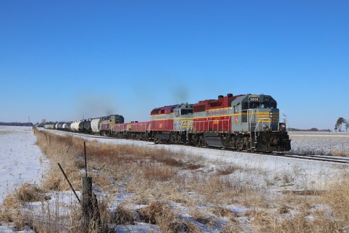 A freight train with colorful locomotives travels through a snowy landscape under a clear blue sky.