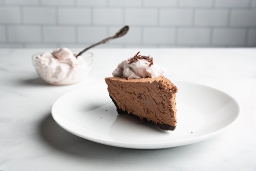 A slice of chocolate mousse pie topped with whipped cream, served on a white plate with a bowl of ice cream in the background.