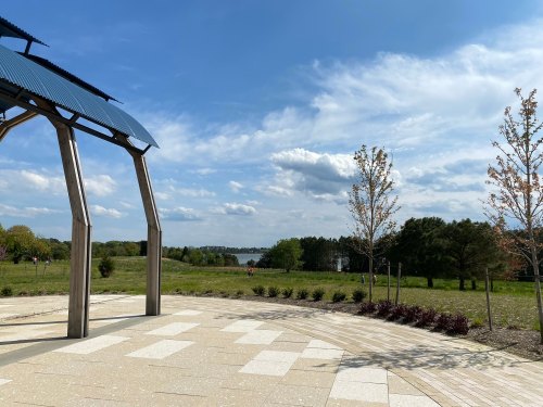 A scenic view of a park with a modern structure, green fields, and a lake under a partly cloudy sky.