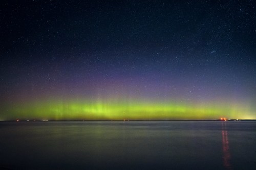 A serene night sky illuminated by vibrant green and purple auroras over a calm body of water, dotted with distant lights.