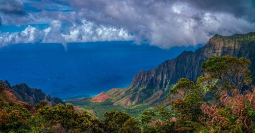 Lush green mountains meet a deep blue ocean under a dramatic sky with clouds, showcasing a stunning natural landscape.