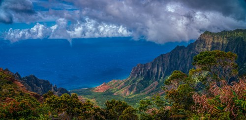 A panoramic view of lush green mountains meeting a deep blue ocean under a dramatic sky with clouds.