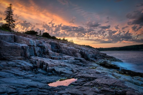 Sunset over rocky coastline with clouds and trees, reflecting in a small pool of water on the rocks.