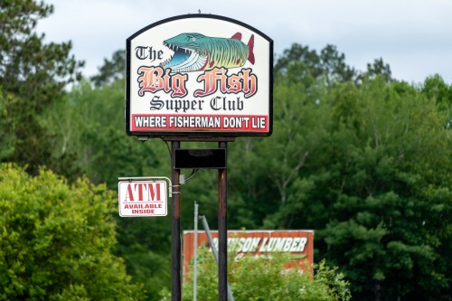 Sign for "The Big Fish Supper Club" featuring a large fish graphic, with a note about an ATM available inside.