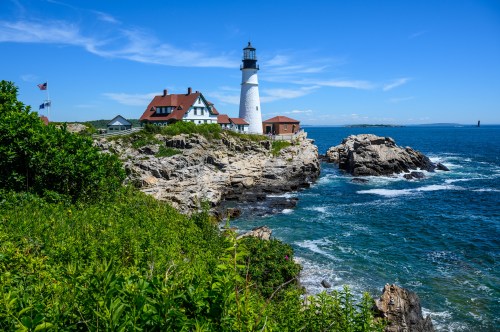 A scenic lighthouse on rocky shores, surrounded by greenery and ocean under a clear blue sky.