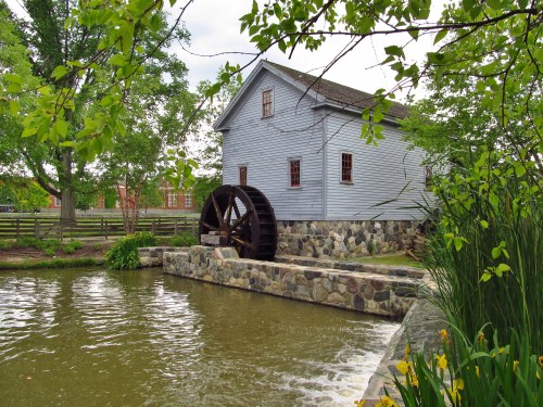 A historic wooden mill with a water wheel beside a calm pond, surrounded by greenery and a stone wall.