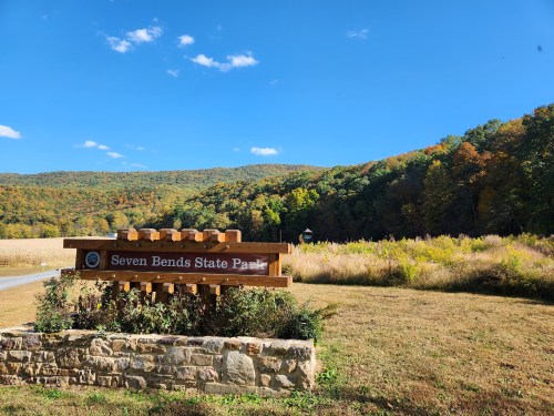 Sign for Seven Bends State Park with colorful autumn trees and a clear blue sky in the background.