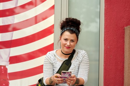 A woman with a bun hairstyle sits outside, smiling while holding a phone, with a red and white striped background.