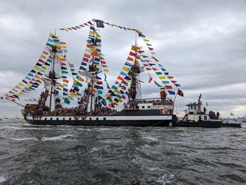A decorated tall ship with colorful flags sails on a cloudy day, accompanied by a small tugboat.