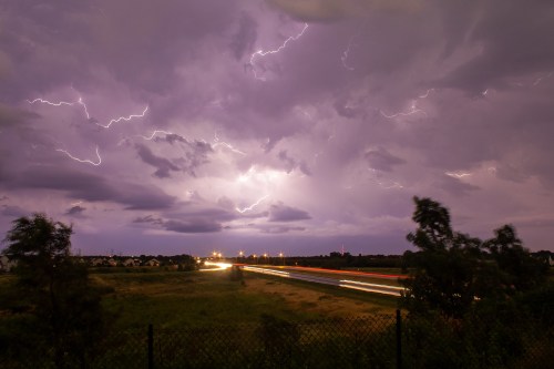 A dramatic sky filled with lightning above a highway, with streaks of car lights and dark, cloudy weather.
