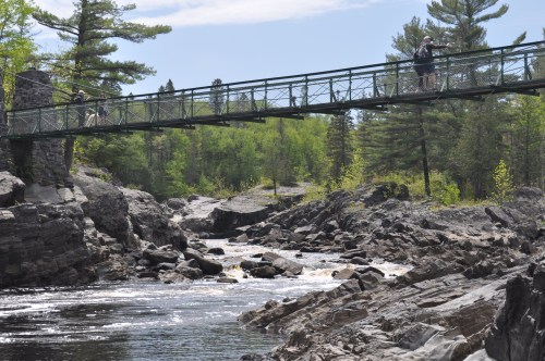 A suspension bridge spans a rocky river, surrounded by lush green trees under a clear blue sky.