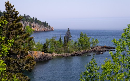 A serene coastal view featuring lush greenery, rocky shores, and calm blue waters under a clear sky.