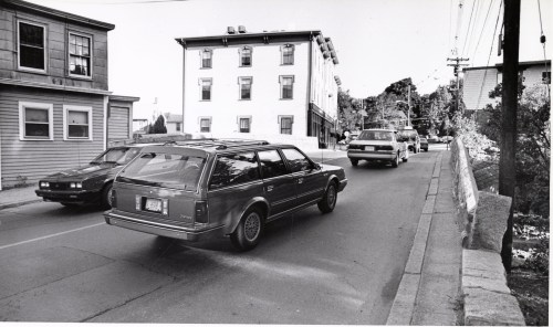 Black and white photo of cars on a street with buildings and trees in the background.